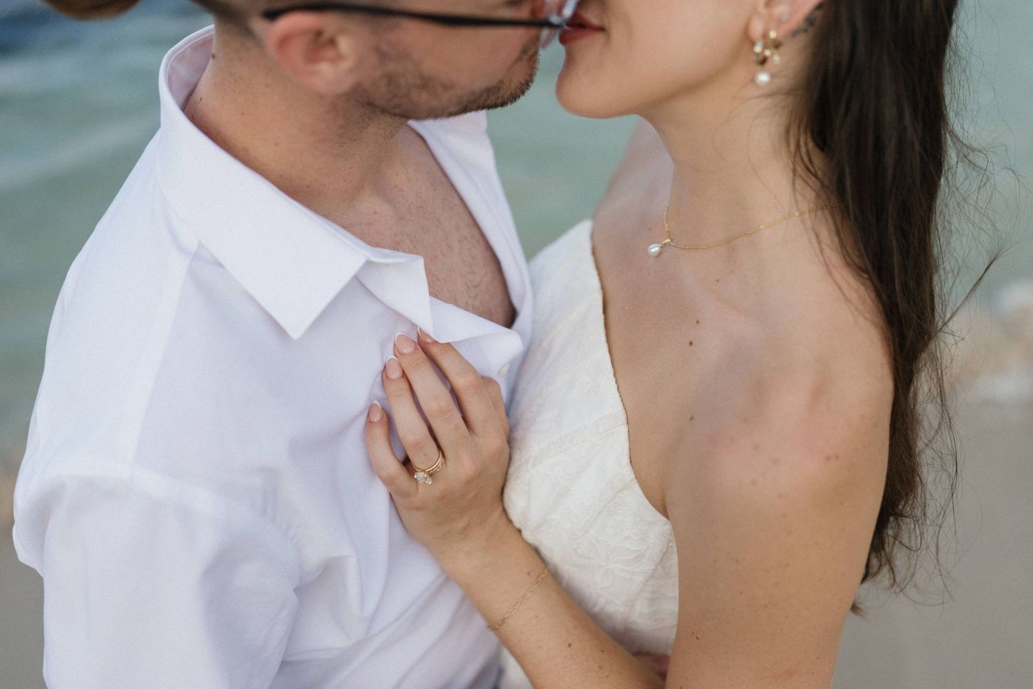 Couple in white attire share a romantic kiss on a beach, her hand resting on his chest, showing a ring, with the ocean blurred behind them.