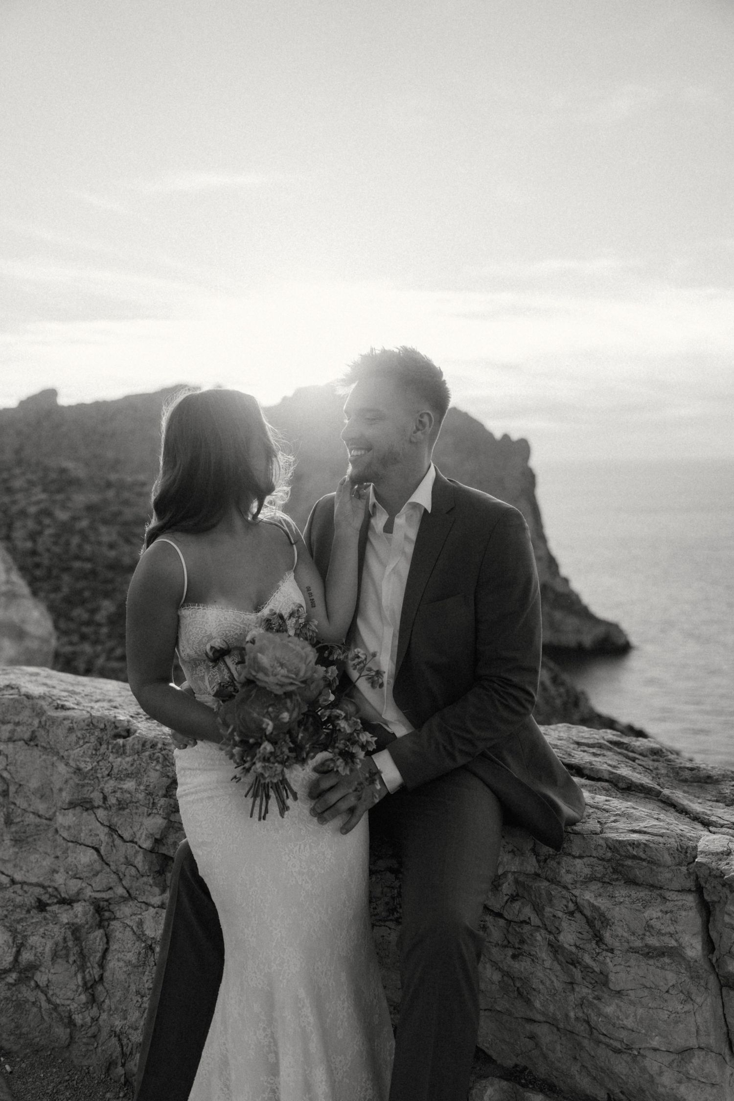 Black and white photo of a bride and groom sharing an intimate moment on rocky cliffs by the ocean at sunset.