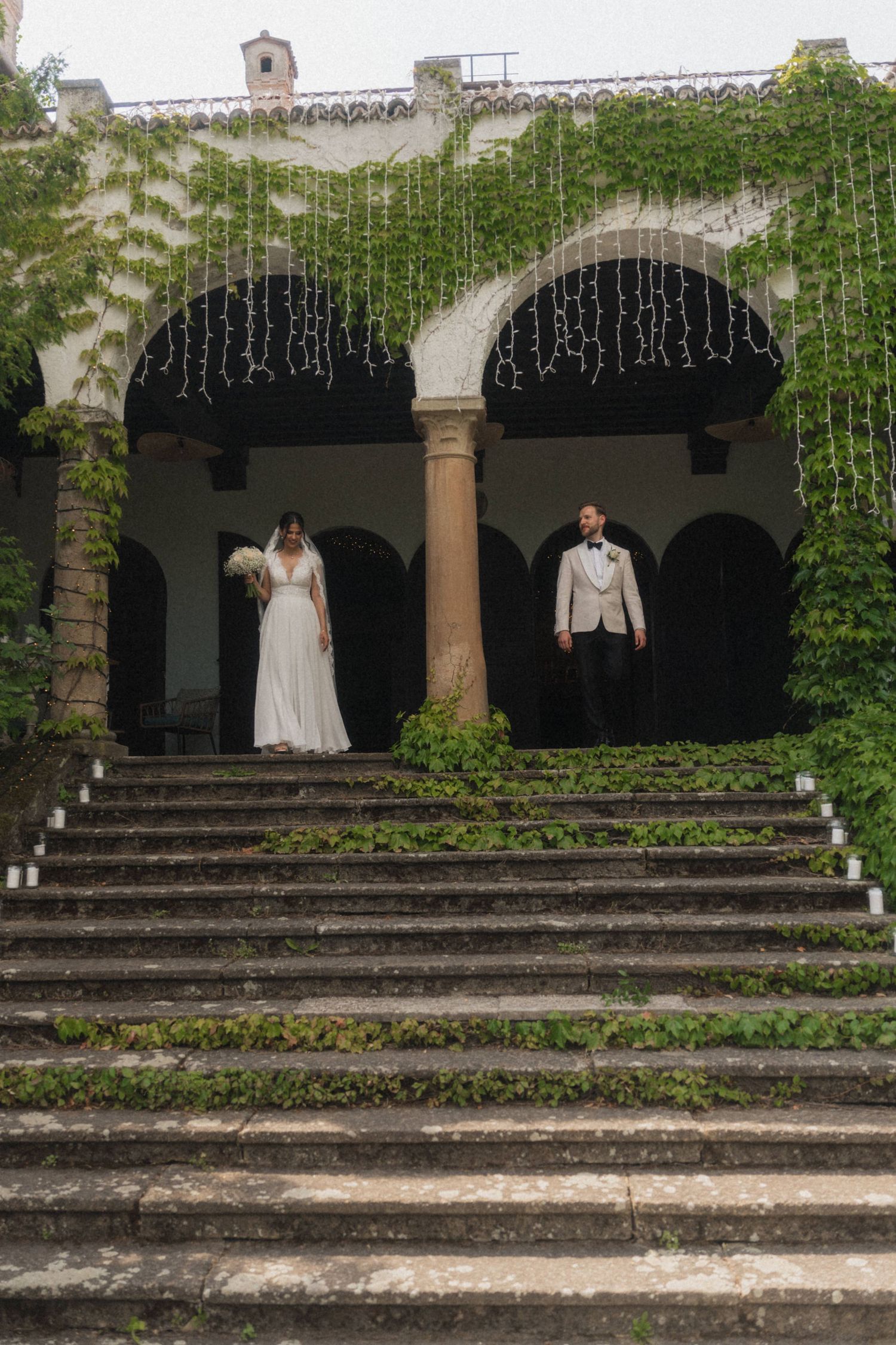 Wedding couple standing under ivy-covered stone arches with string lights on moss-covered steps.