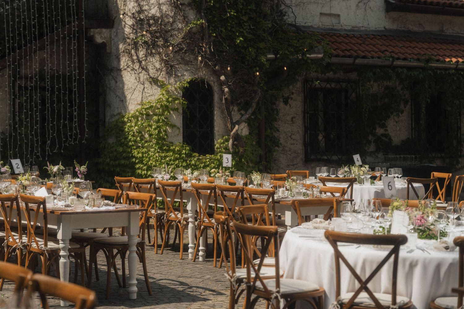 Sunlit outdoor wedding reception courtyard with wooden cross-back chairs and elegantly set round tables.