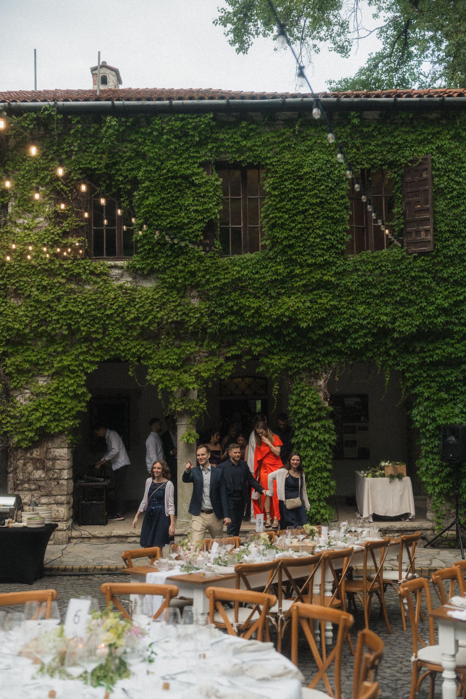 Wedding guests gather at outdoor reception tables before ivy-covered stone villa with string lights.