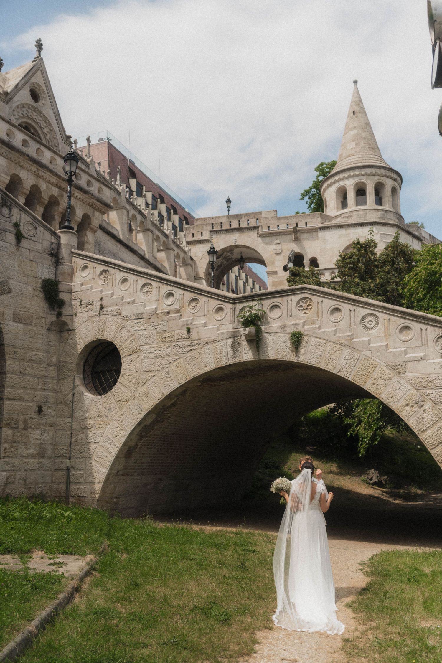 Bride in white gown and veil stands before a historic stone bridge and castle tower in Budapest.