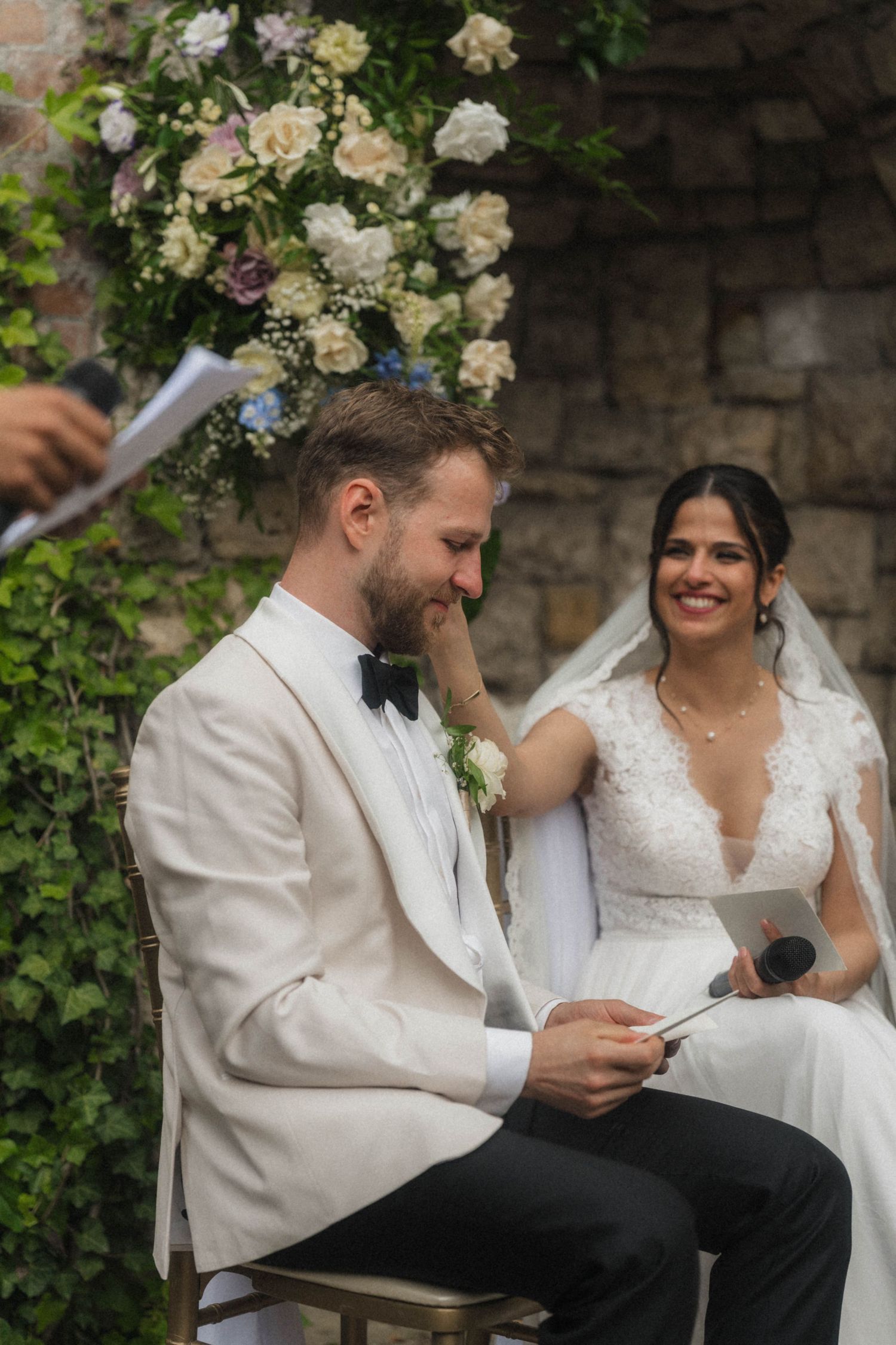 Bride and groom seated during outdoor wedding ceremony, groom in white tuxedo, bride in lace gown smiling.