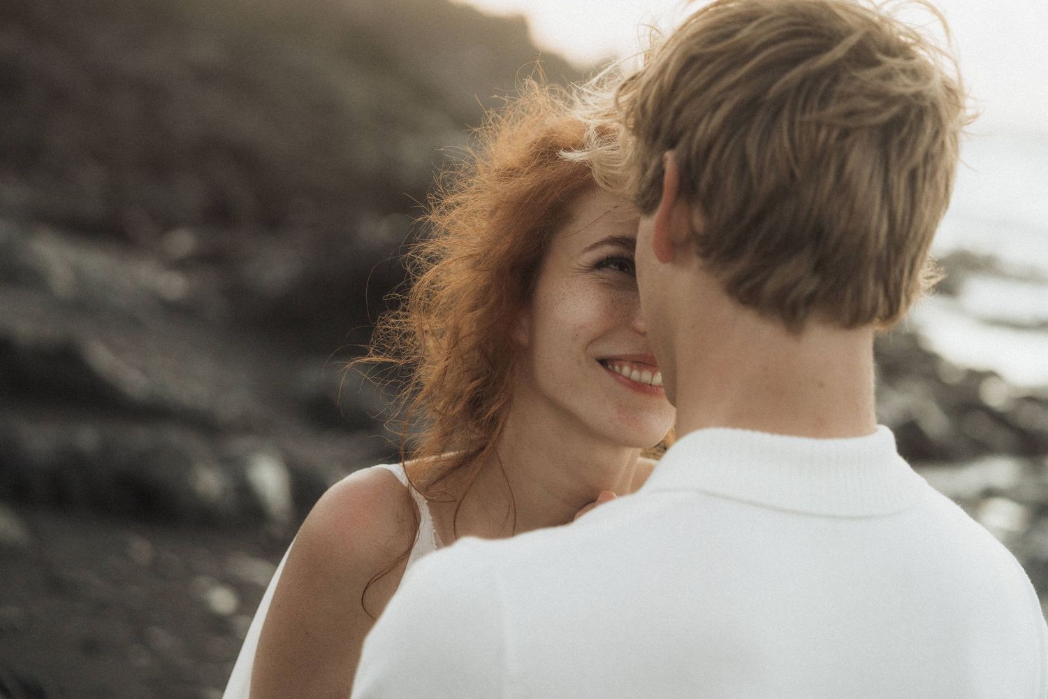 Smiling redhead bride gazes at blonde groom on rocky beach at golden hour.