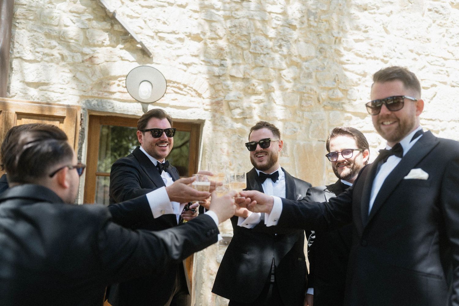 Groomsmen in black tuxedos and sunglasses toasting drinks outside a stone building.