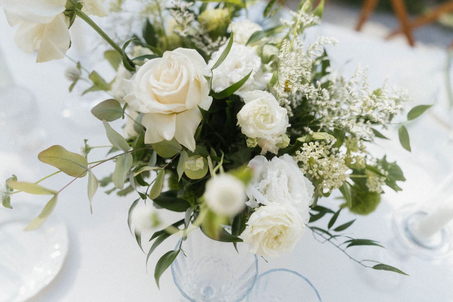 Elegant white rose and greenery wedding centerpiece arrangement in a glass vase on a white tablecloth.