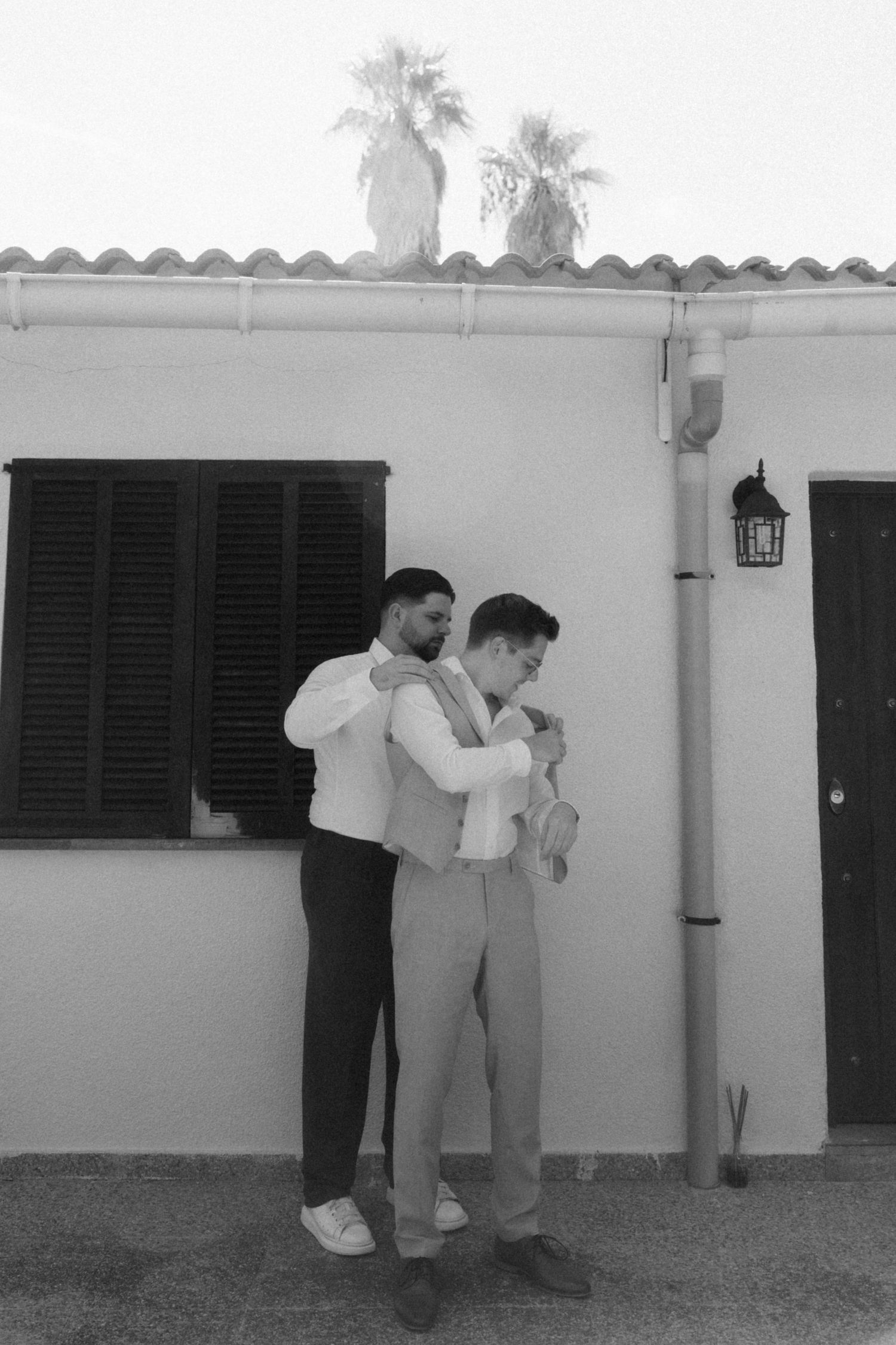 Black and white photo of two men in suits helping each other get dressed outside building.