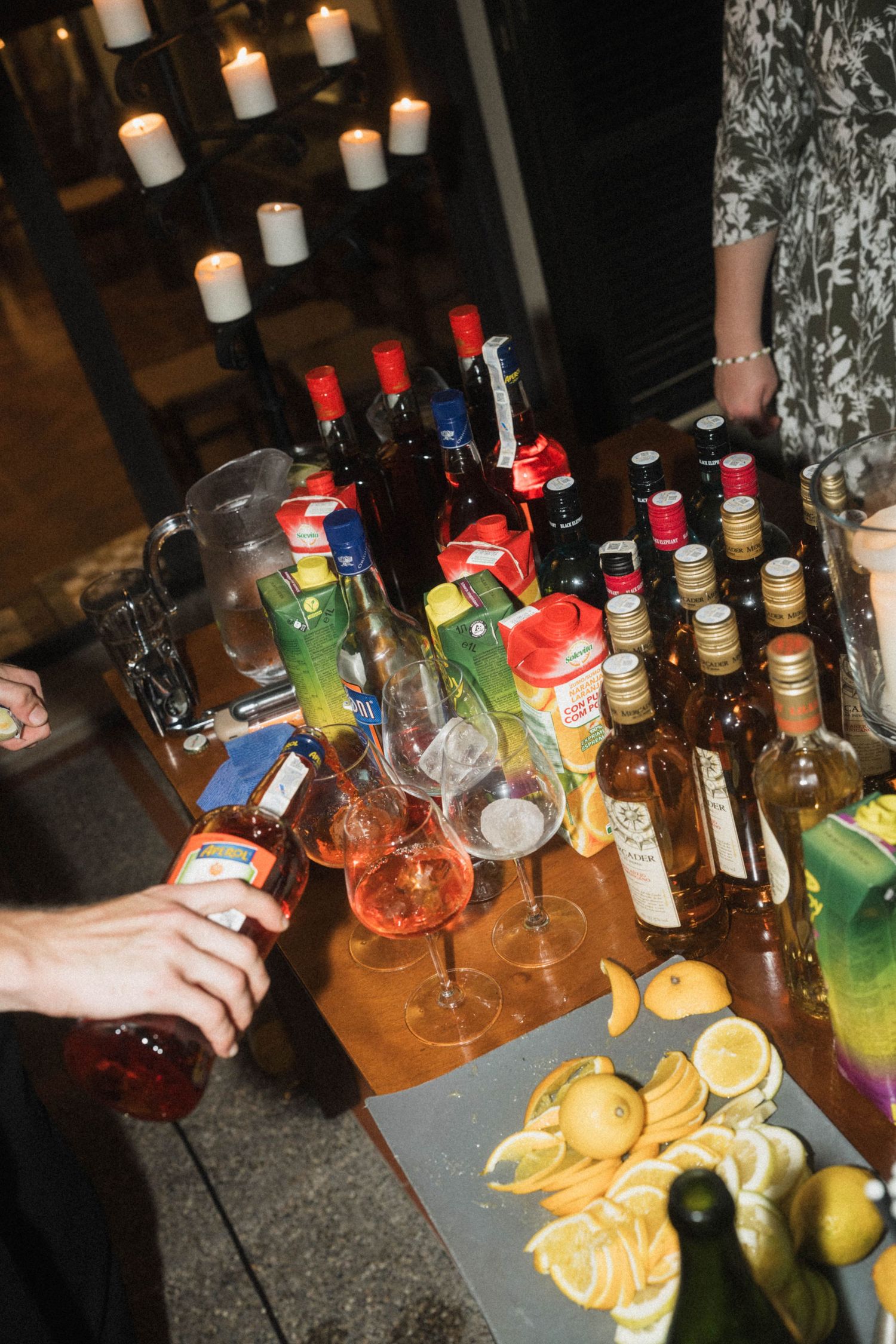 Bartender pouring cocktails at a bar setup with assorted liquor bottles, wine glasses, and fresh citrus fruits.