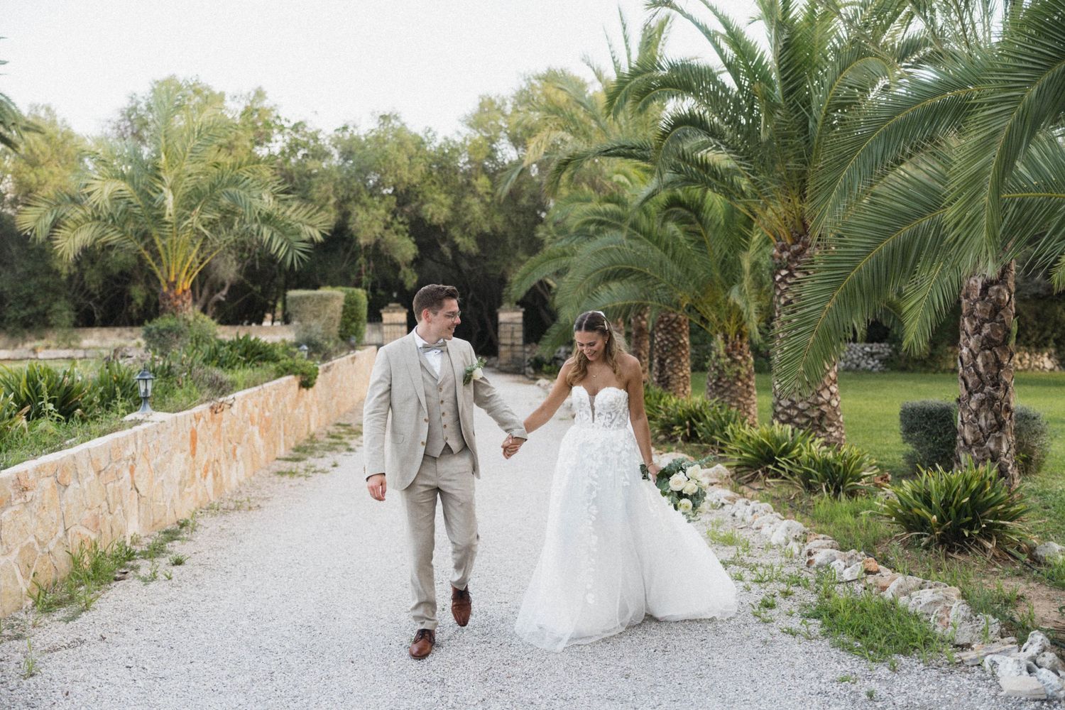 Bride and groom holding hands walking along a palm-lined gravel path at an outdoor wedding venue.