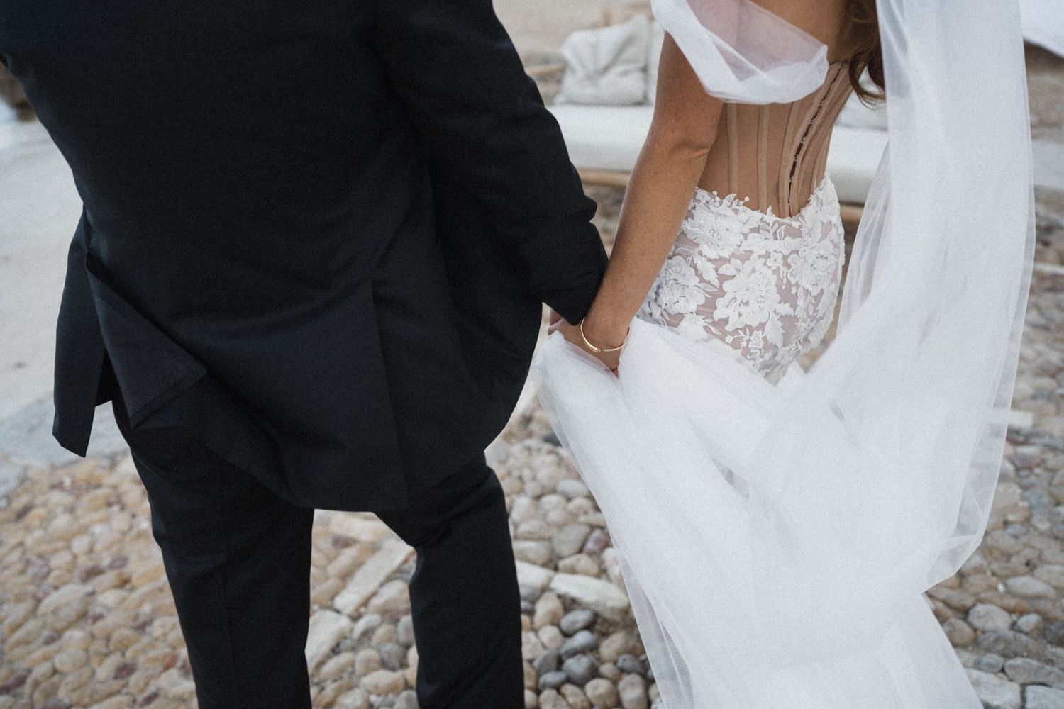 Close-up of bride in lace gown holding groom's hand on pebbled path.