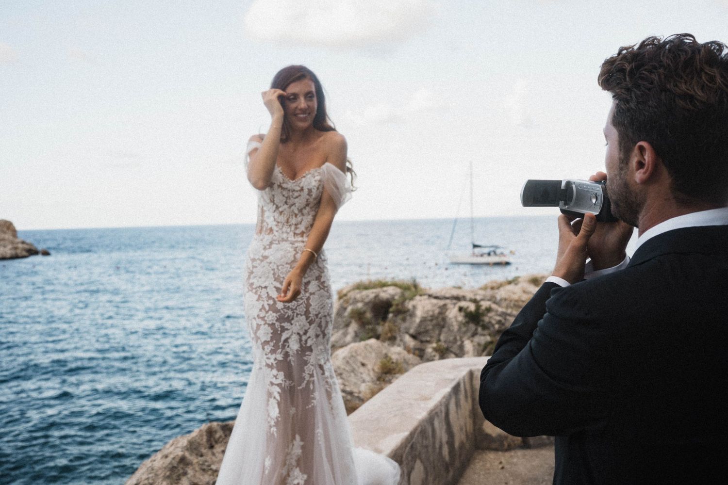 Groom capturing bride posing in floral lace mermaid wedding dress on rocky coastal cliff by the sea.