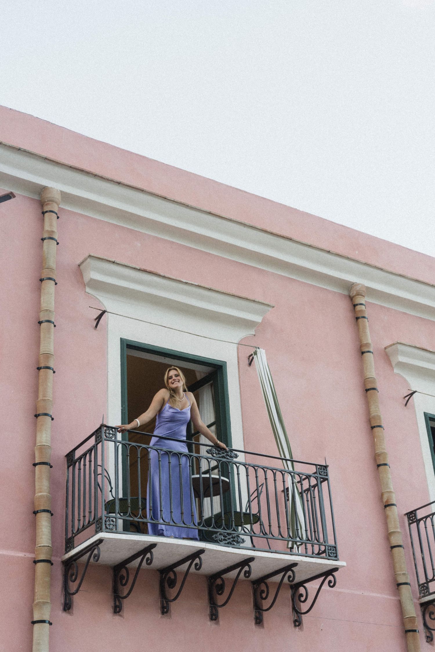Bridesmaid in lavender gown stands on balcony of a pink building at a destination wedding.