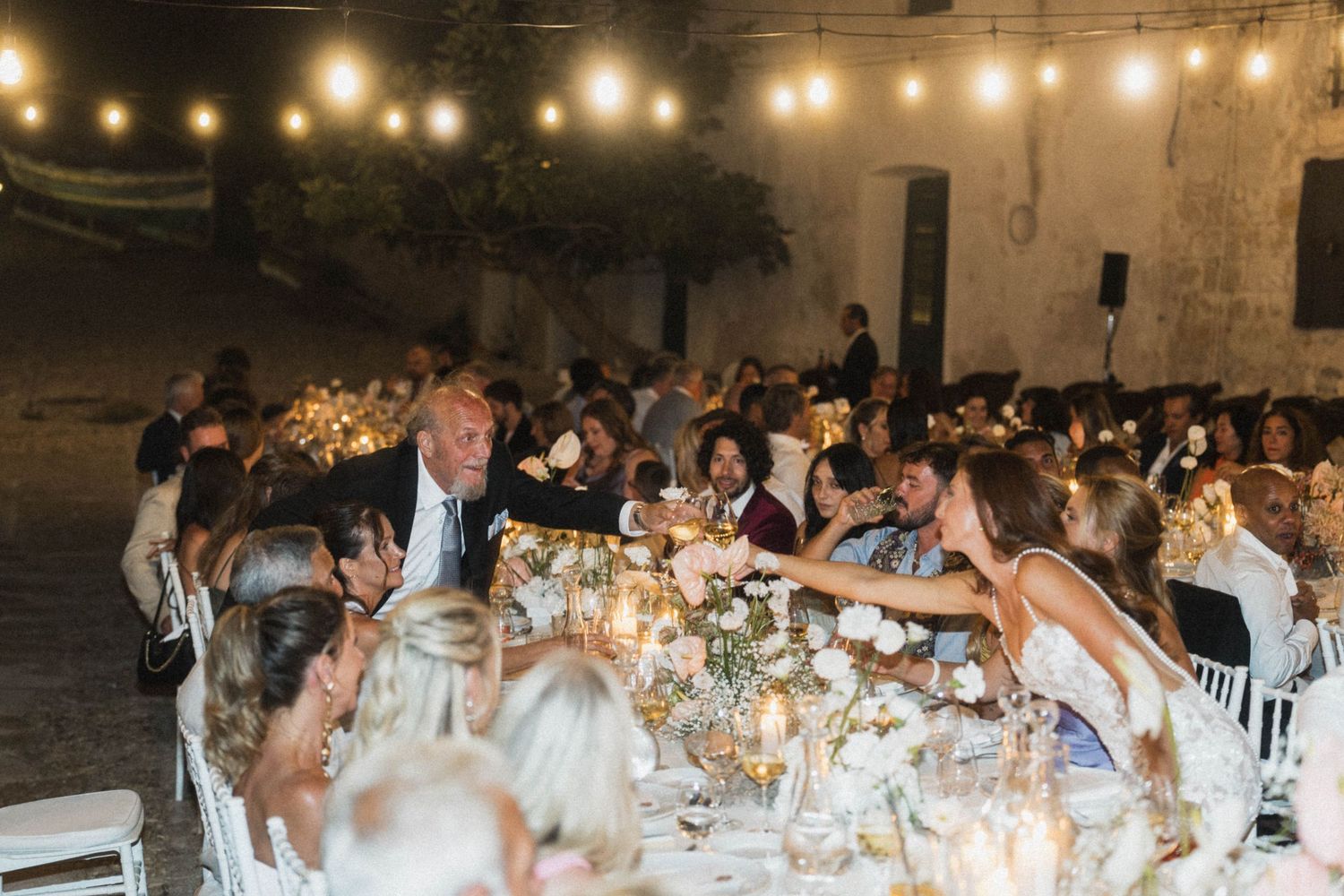 Wedding guests raising glasses in a toast at an elegant candlelit outdoor reception with string lights overhead.