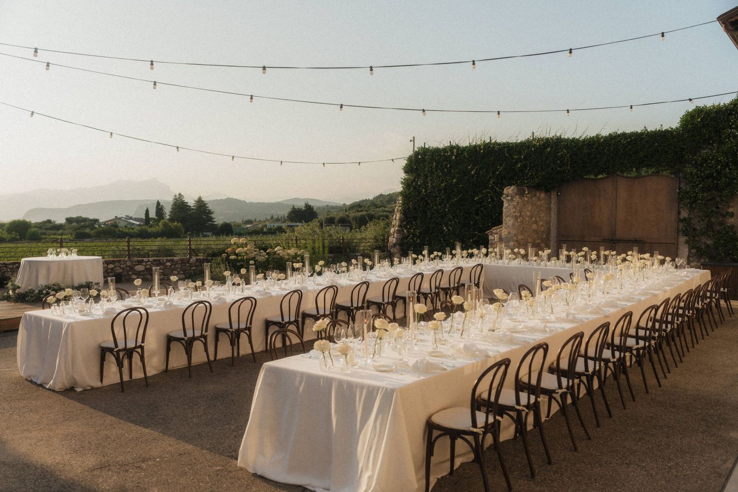 Elegant outdoor wedding reception with long white tables, string lights, and mountain views.