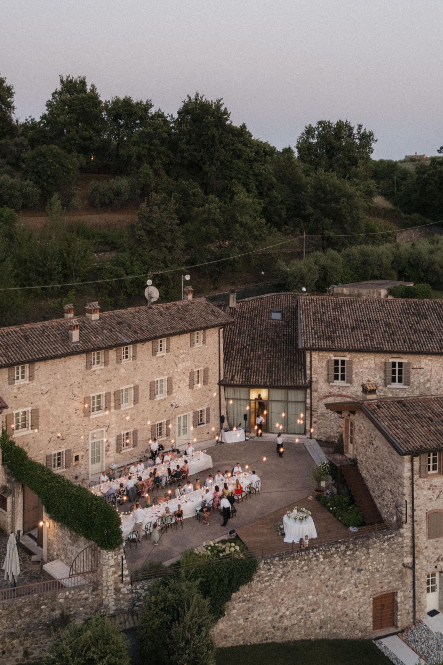 Aerial twilight view of rustic Italian villa wedding reception with guests dining outdoors.