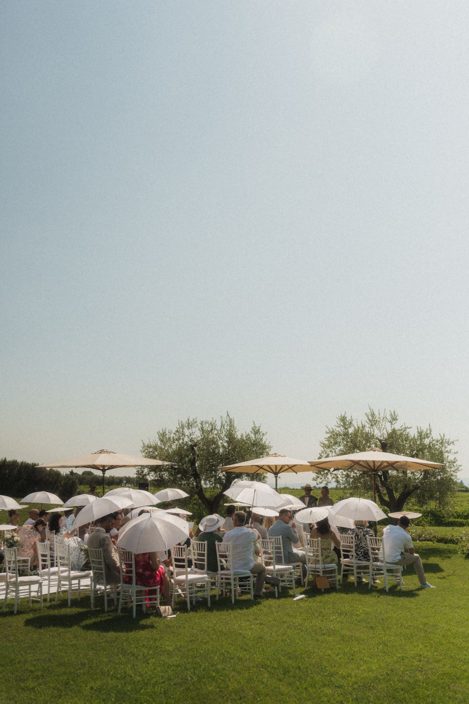 Outdoor wedding ceremony on green lawn with guests seated on white chairs under white parasols on a sunny day.