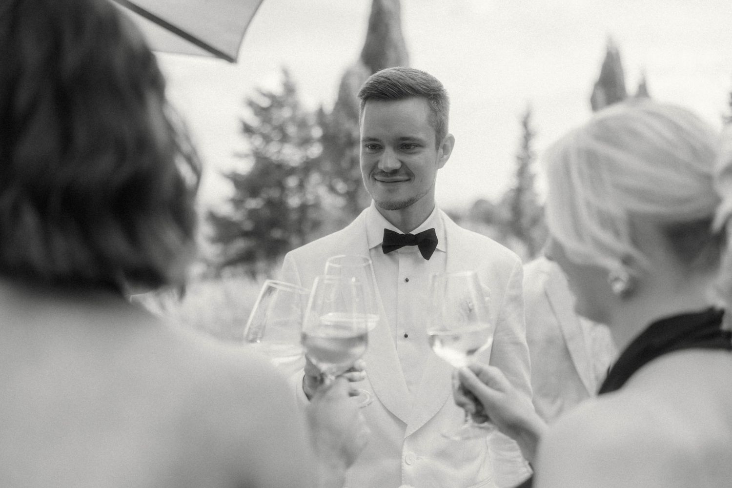 Young man in white tuxedo and bow tie holding wine glass at elegant outdoor event, black and white photo.