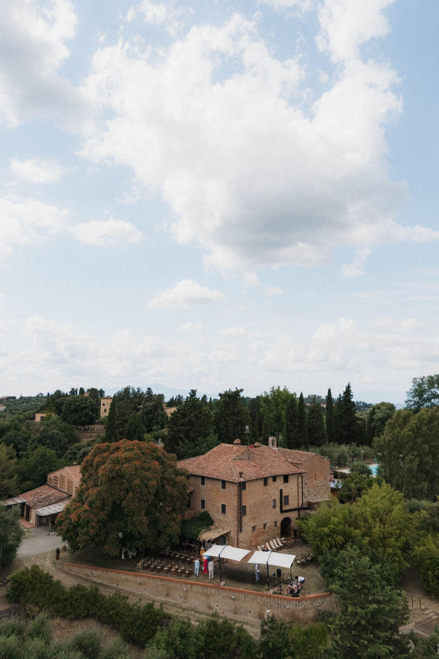 Aerial view of a rustic Tuscan villa surrounded by lush green trees under a cloudy sky.