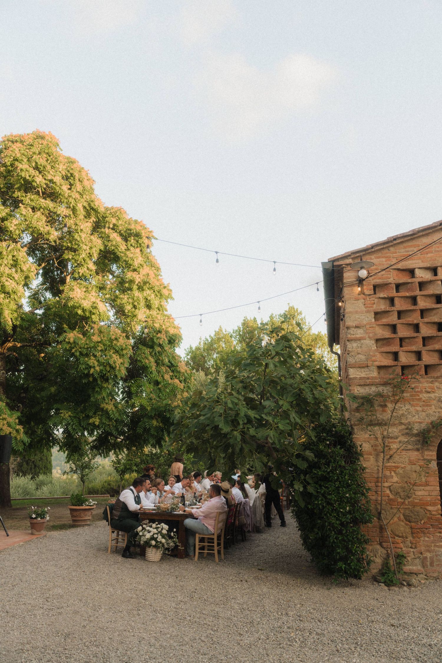 Wedding guests dining at outdoor long table beside rustic brick building with string lights at dusk.