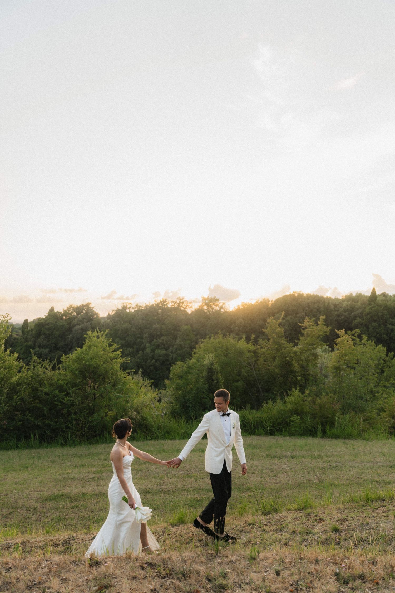 Bride and groom holding hands walking through a sunlit field at golden hour with lush green trees in the background.