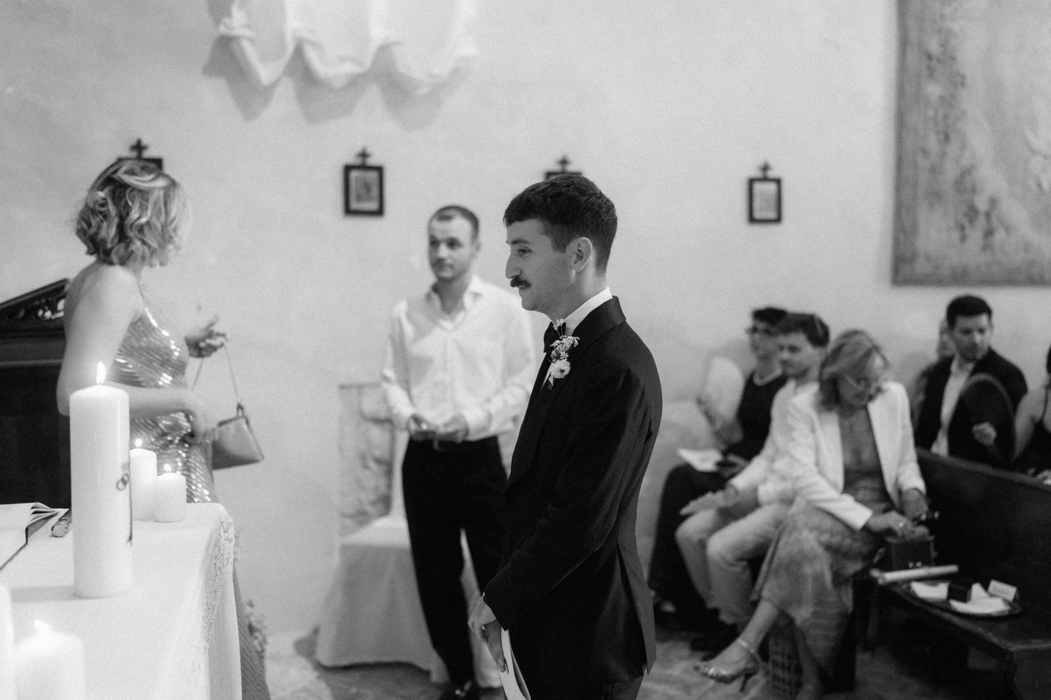 Black and white wedding ceremony photo showing groom in suit standing at altar with officiant and seated guests in background.