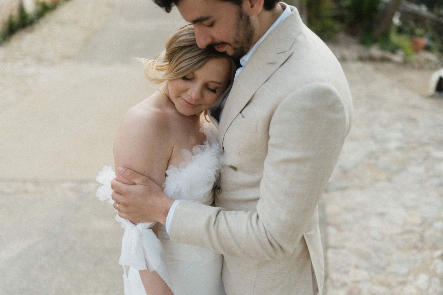 Groom in beige suit tenderly embracing smiling bride in white strapless wedding dress outdoors.