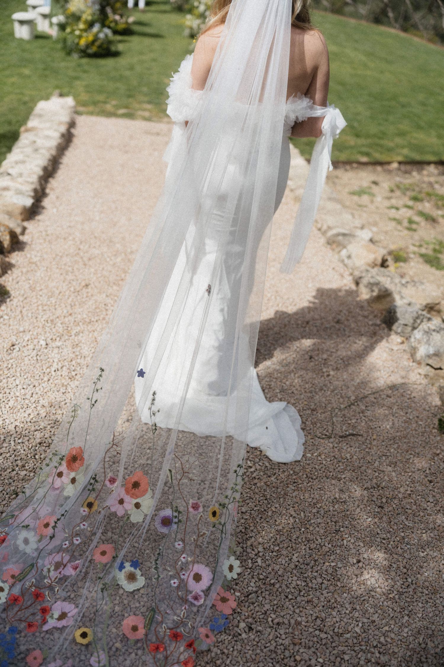 Bride in white wedding dress with floral embroidered veil trailing on stone path in garden.