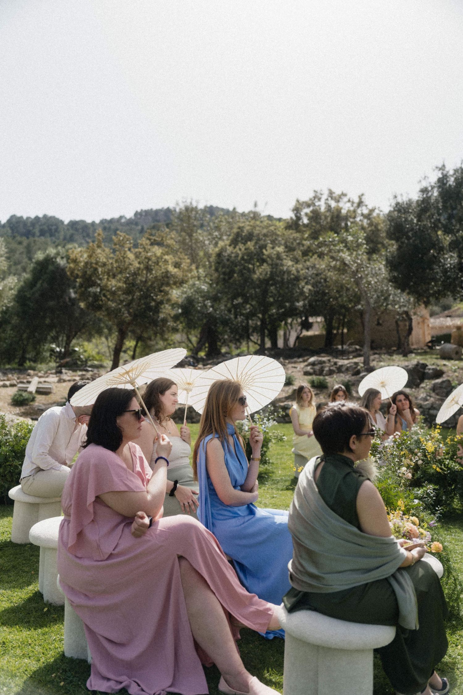 Wedding guests seated outdoors holding white parasols at a sunny garden ceremony with mountain backdrop.