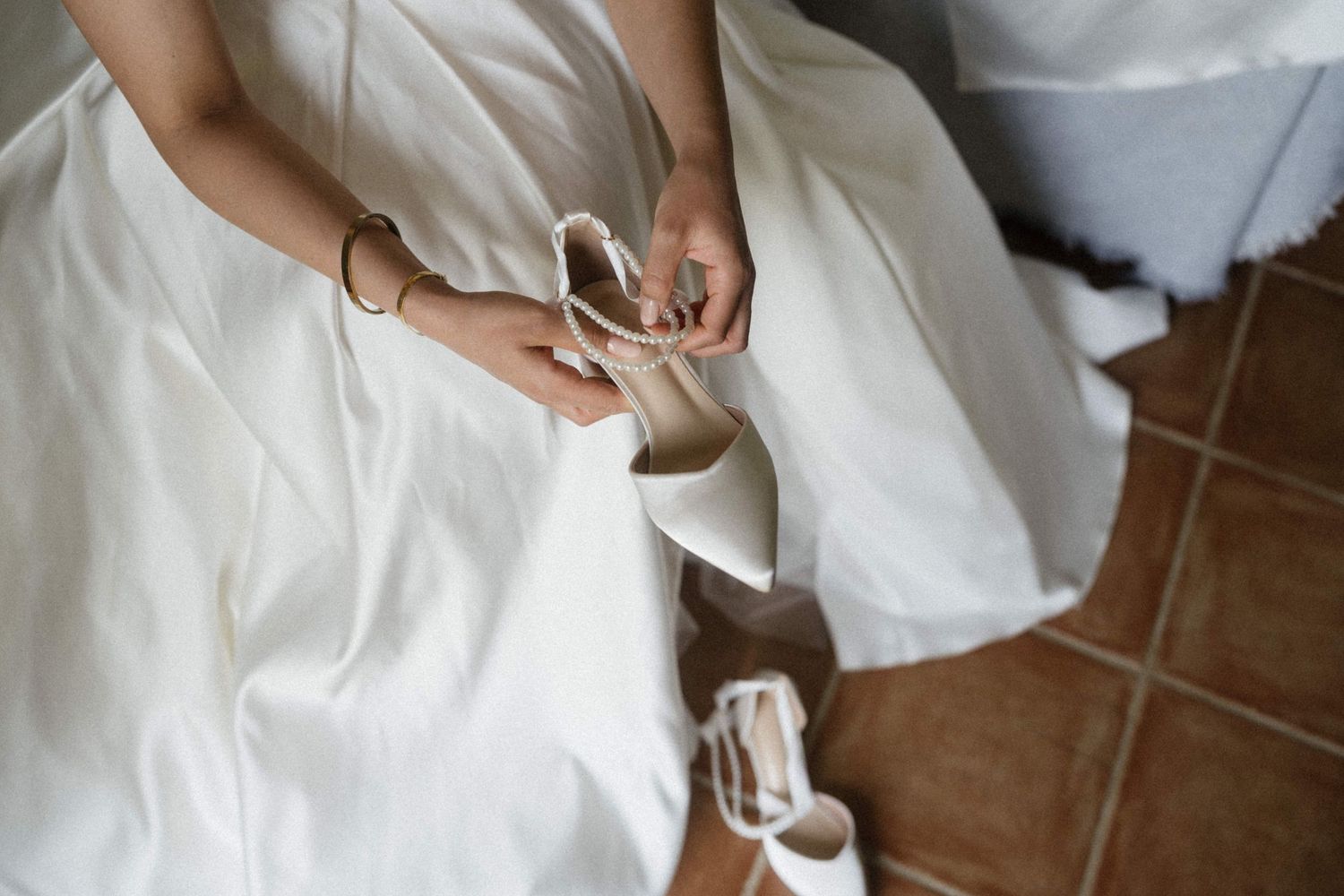 Bride in white wedding dress tying straps on elegant ivory heeled bridal shoes while seated on tiled floor.