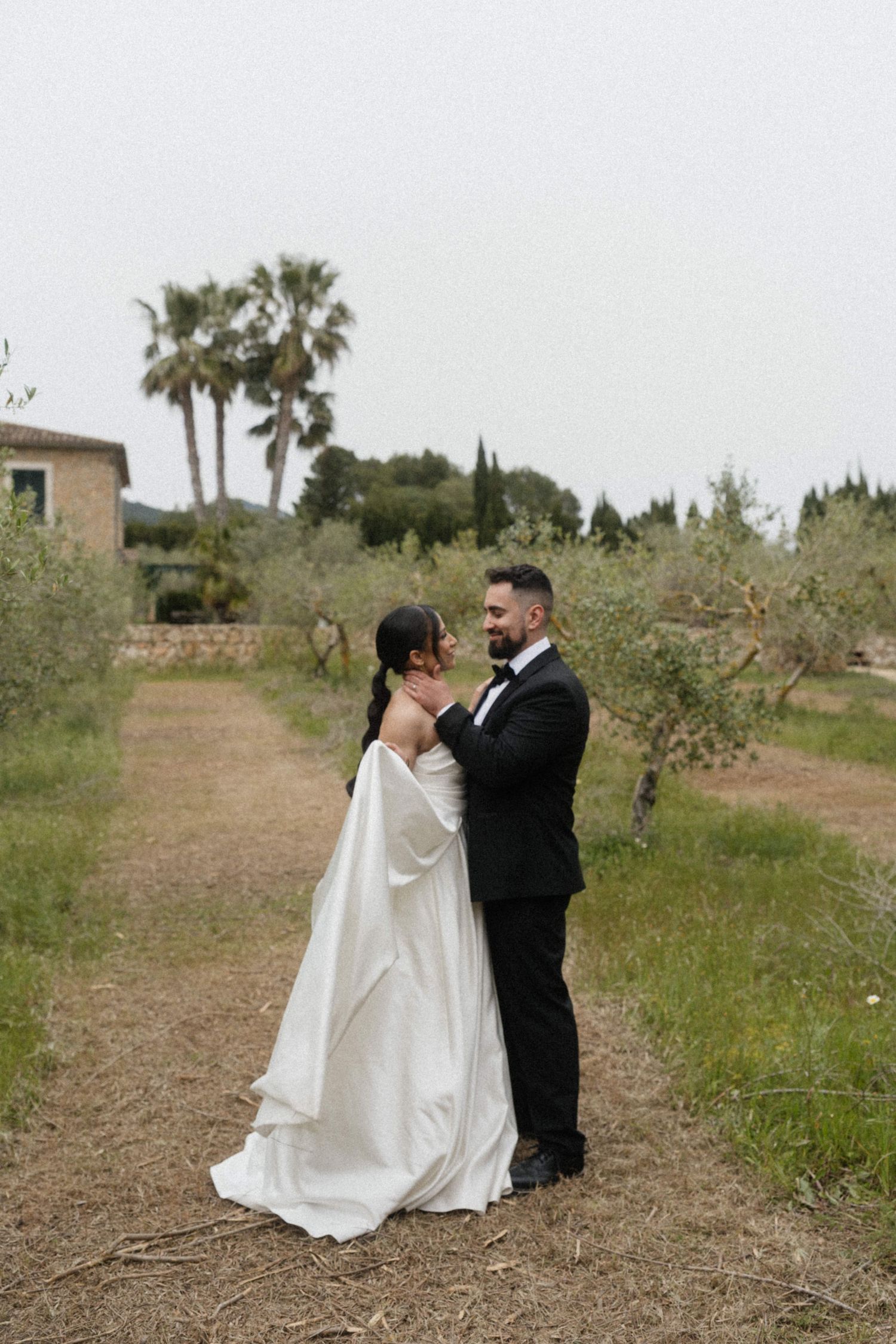 Bride in white cape gown and groom in black tuxedo share a romantic moment in a Mediterranean garden.