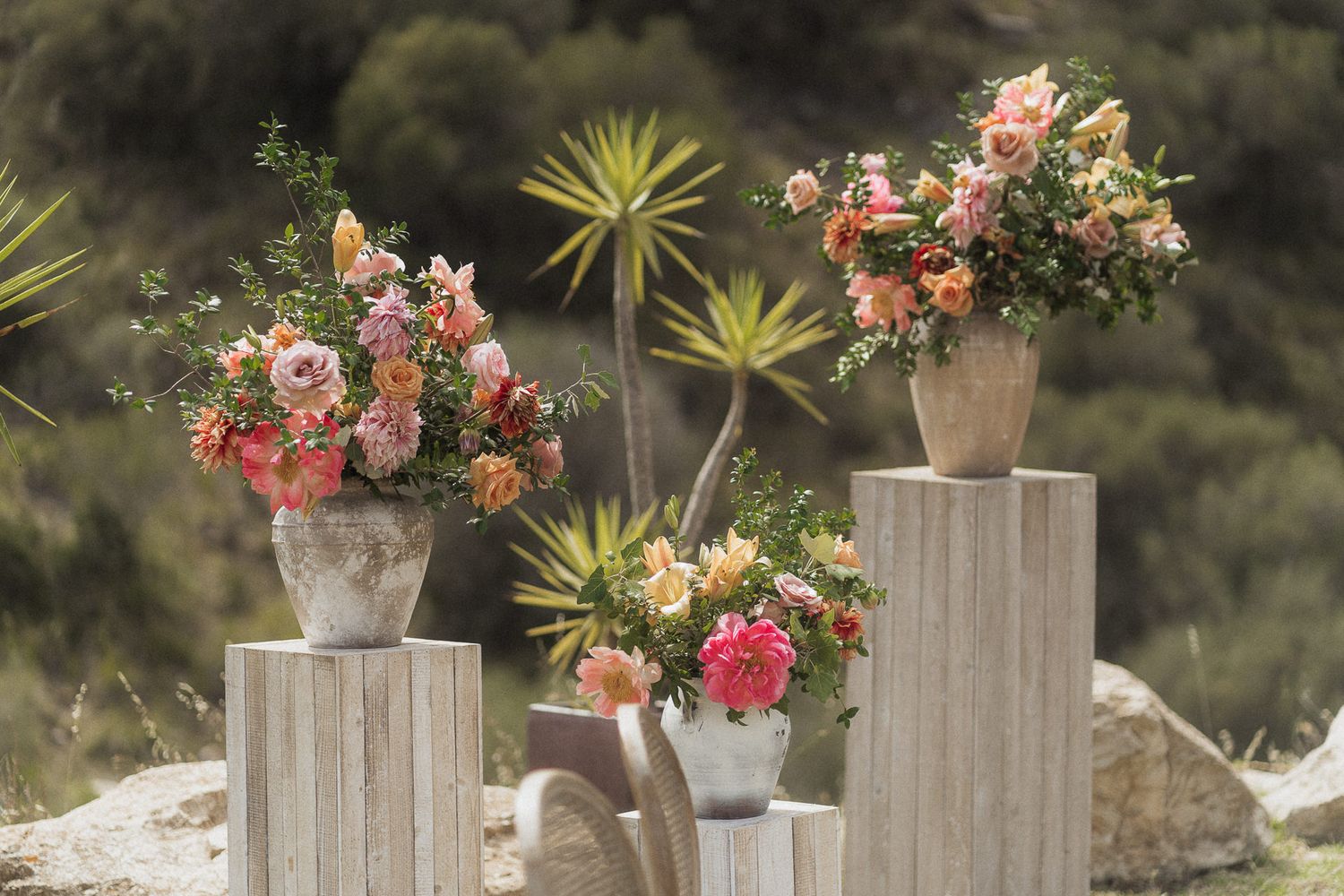 Four rustic stone vases with lush pink and peach floral arrangements displayed on wooden pedestals outdoors.