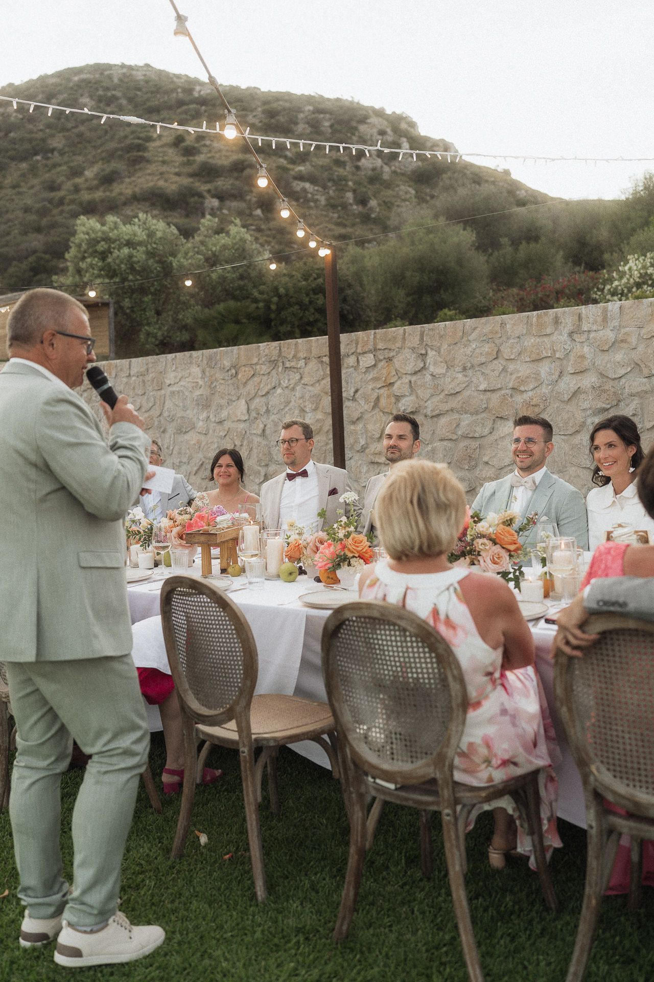 Outdoor wedding reception dinner with guests seated at elegant table, string lights, and stone wall backdrop.