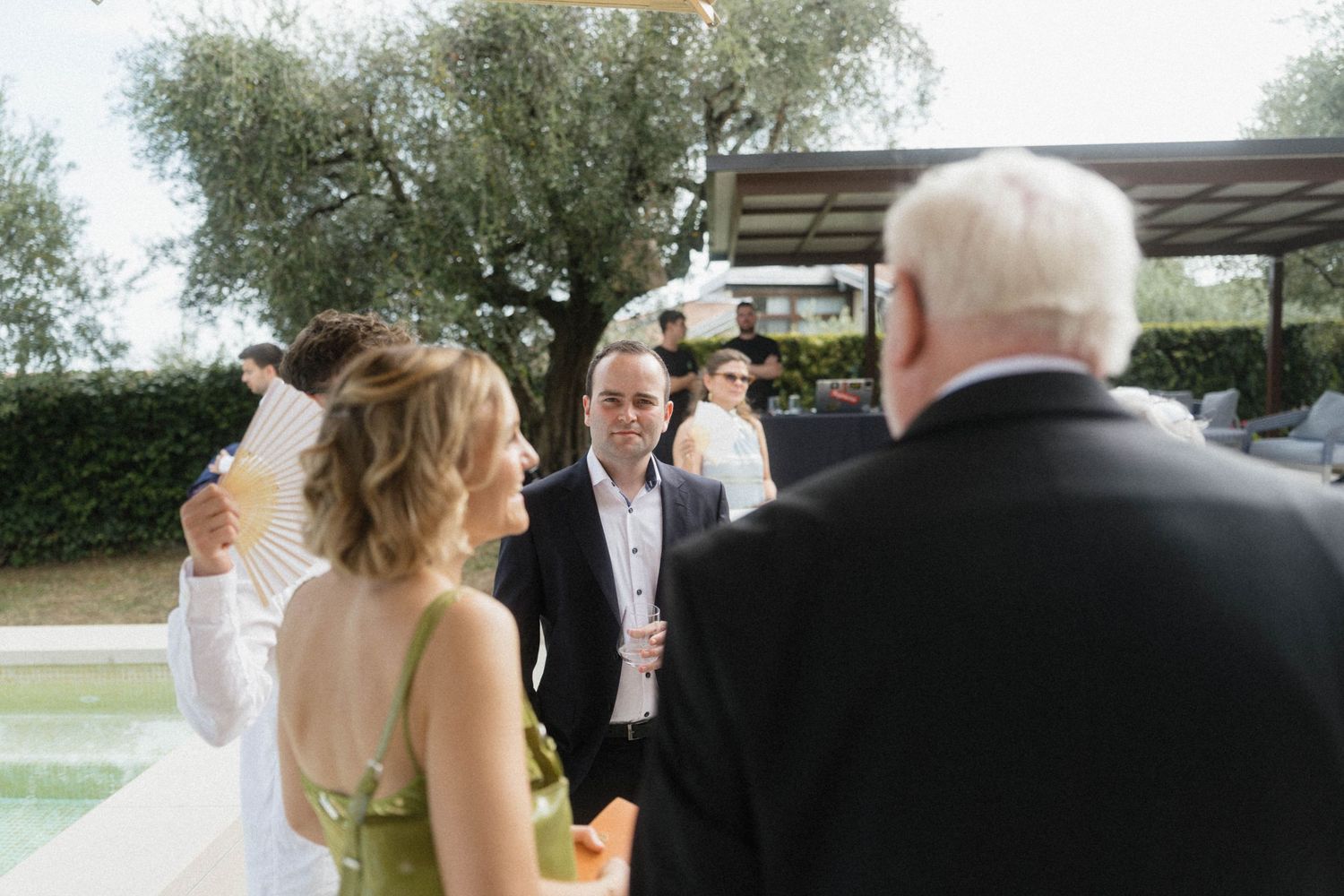 Guests socializing outdoors at a wedding reception near a pool, with a woman holding a decorative fan.