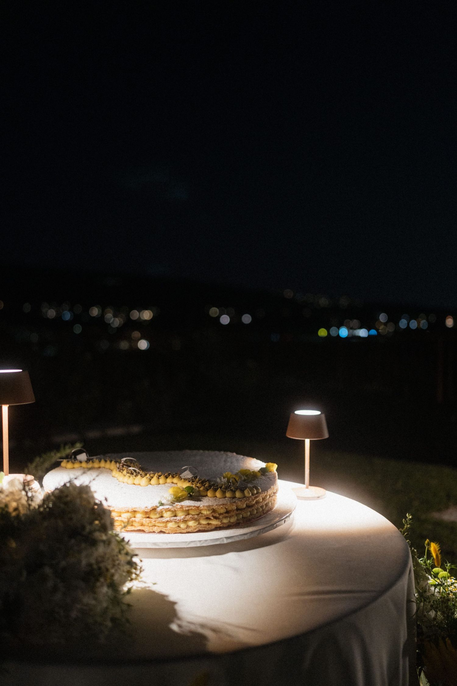 Elegant wedding cake illuminated by small table lamps on a white draped table at night with city lights in background.