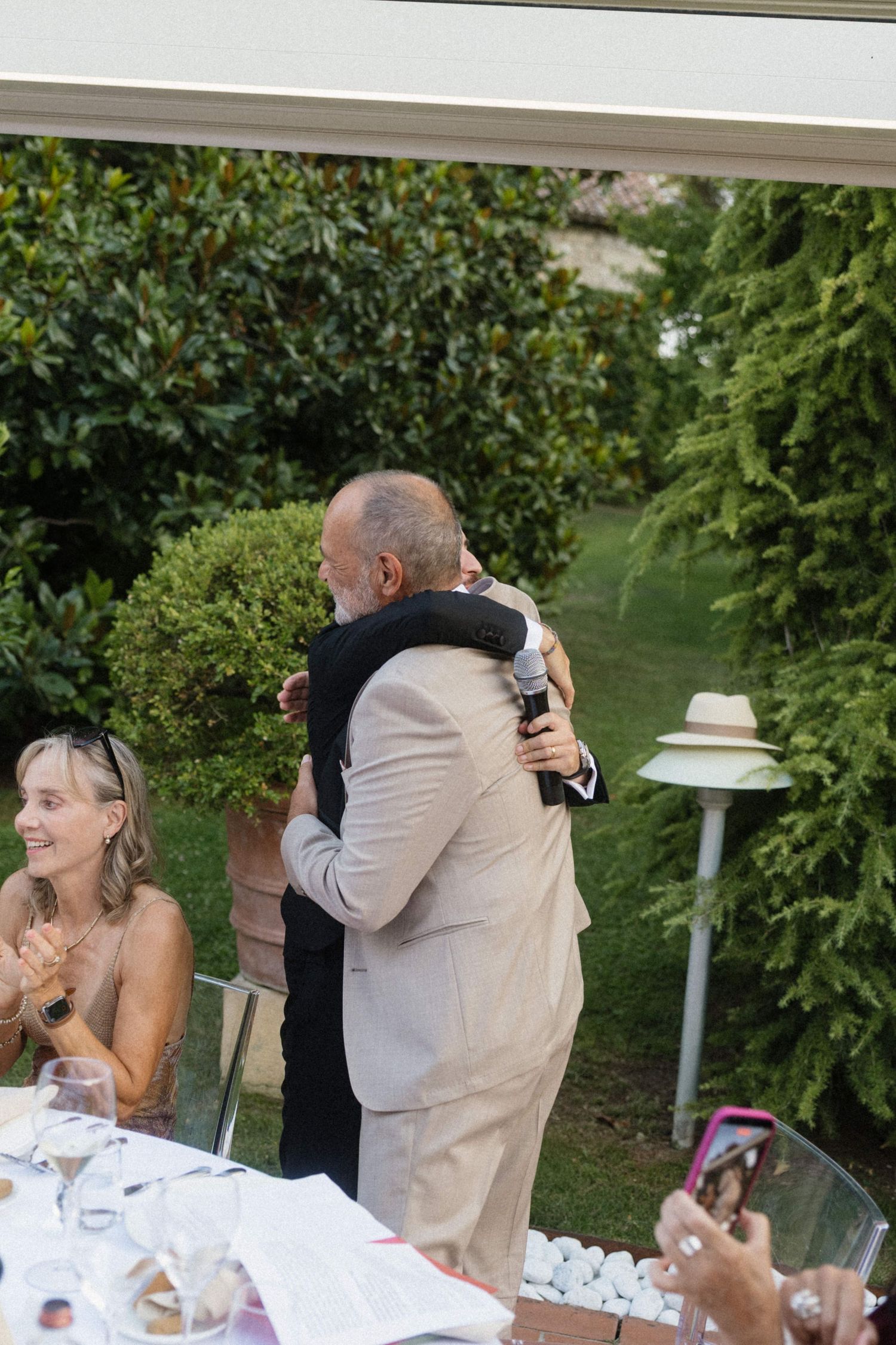 Two men sharing a warm hug during outdoor wedding reception speech while guests applaud and smile.