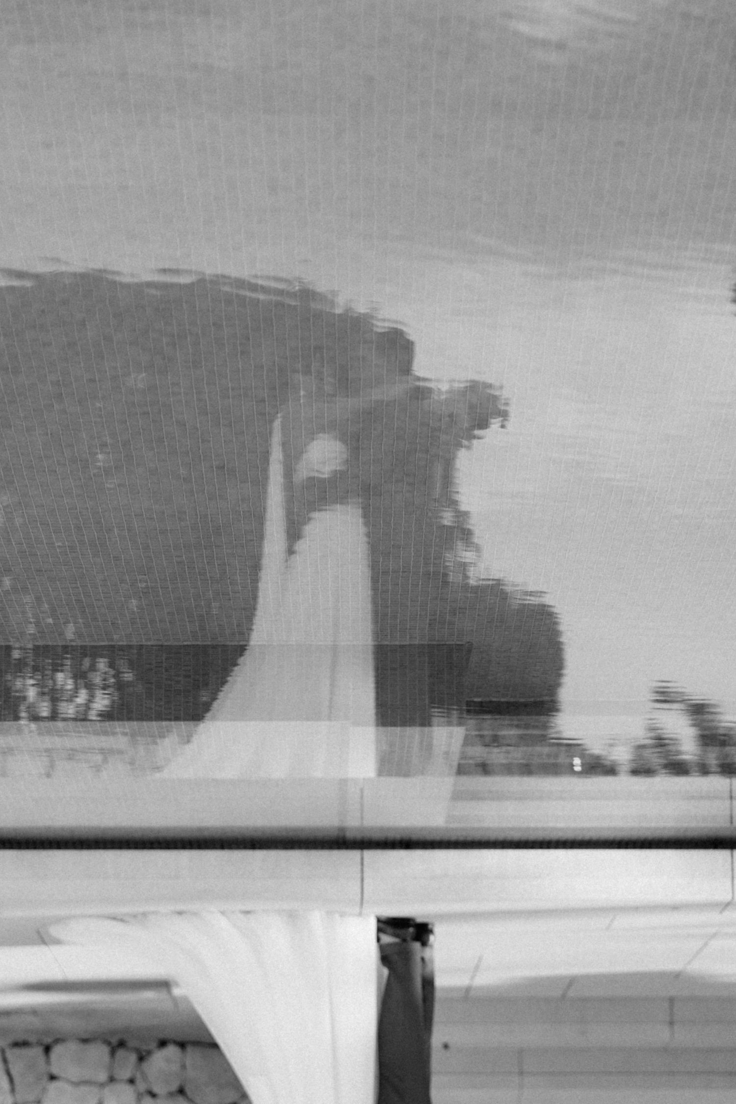 Black and white reflection of a bride in a white gown captured through a window screen on her wedding day.