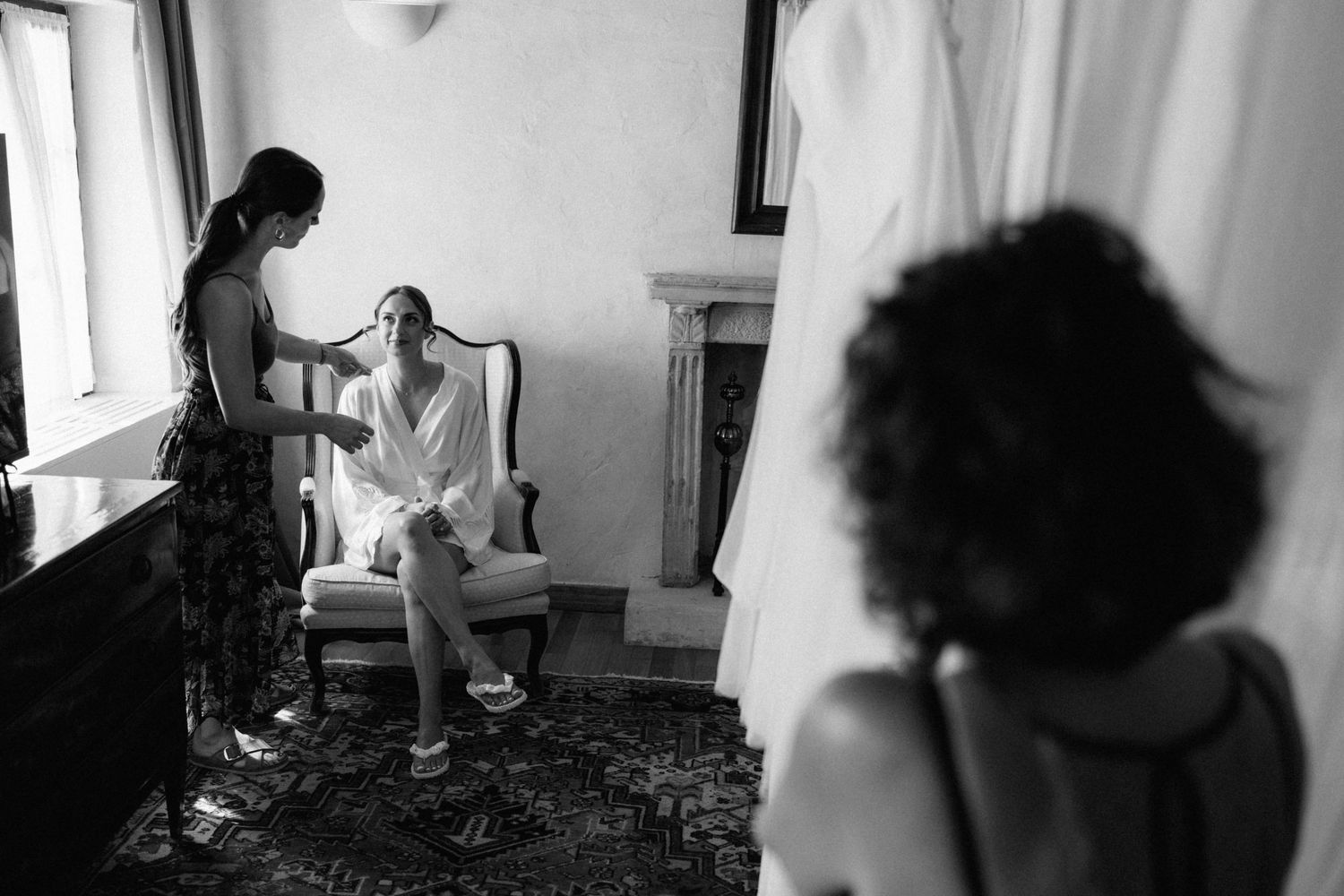 Black and white photo of bride in white robe getting hair done by bridesmaid in elegant room with fireplace.