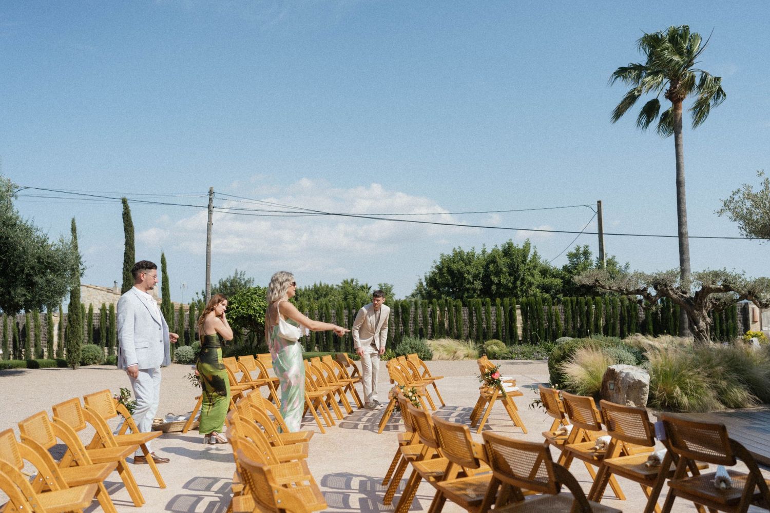 Outdoor wedding ceremony setup with wooden chairs arranged in rows, bridal party walking down the aisle under a sunny blue sky.