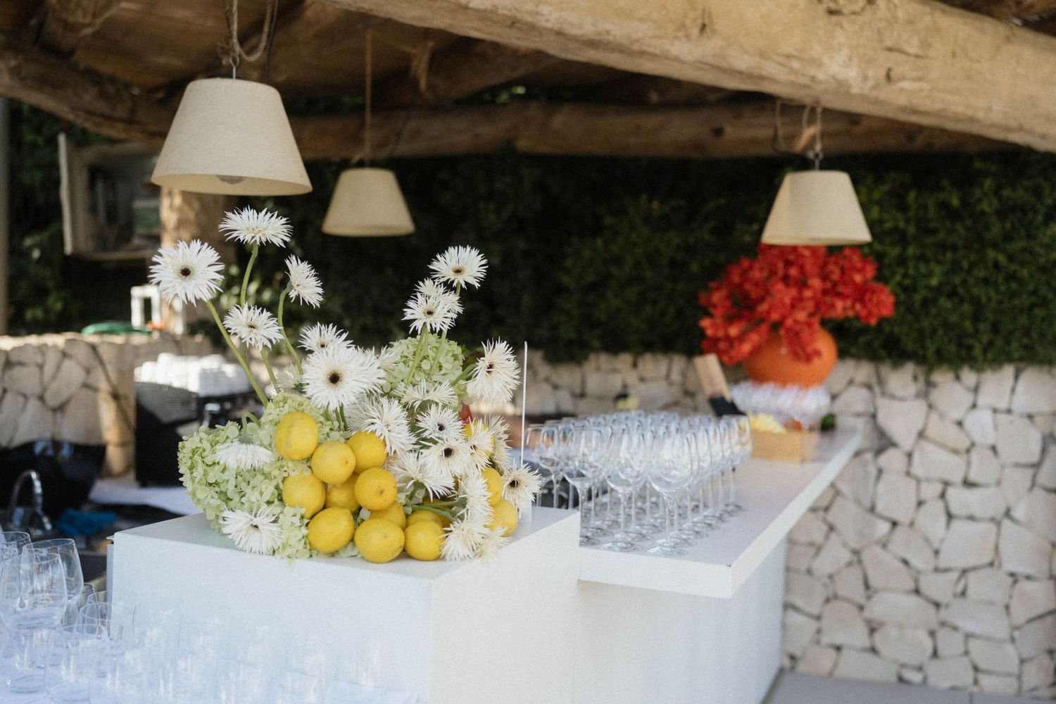 Elegant outdoor bar setup with white flowers, lemons, glassware, and hanging pendant lights at an event.