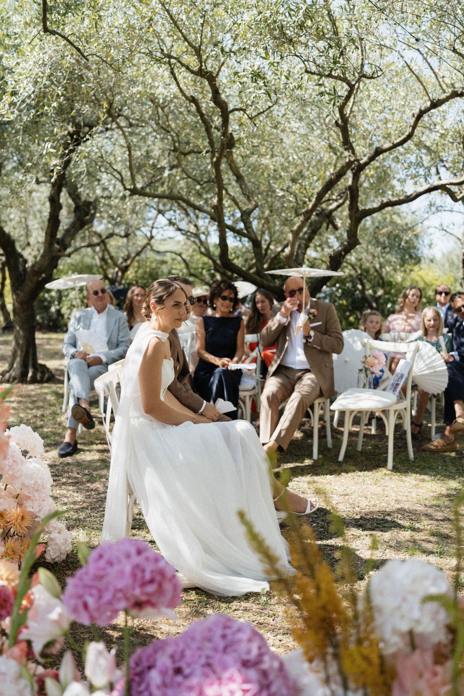 Bride in white gown sits among guests during an outdoor wedding ceremony under olive trees.