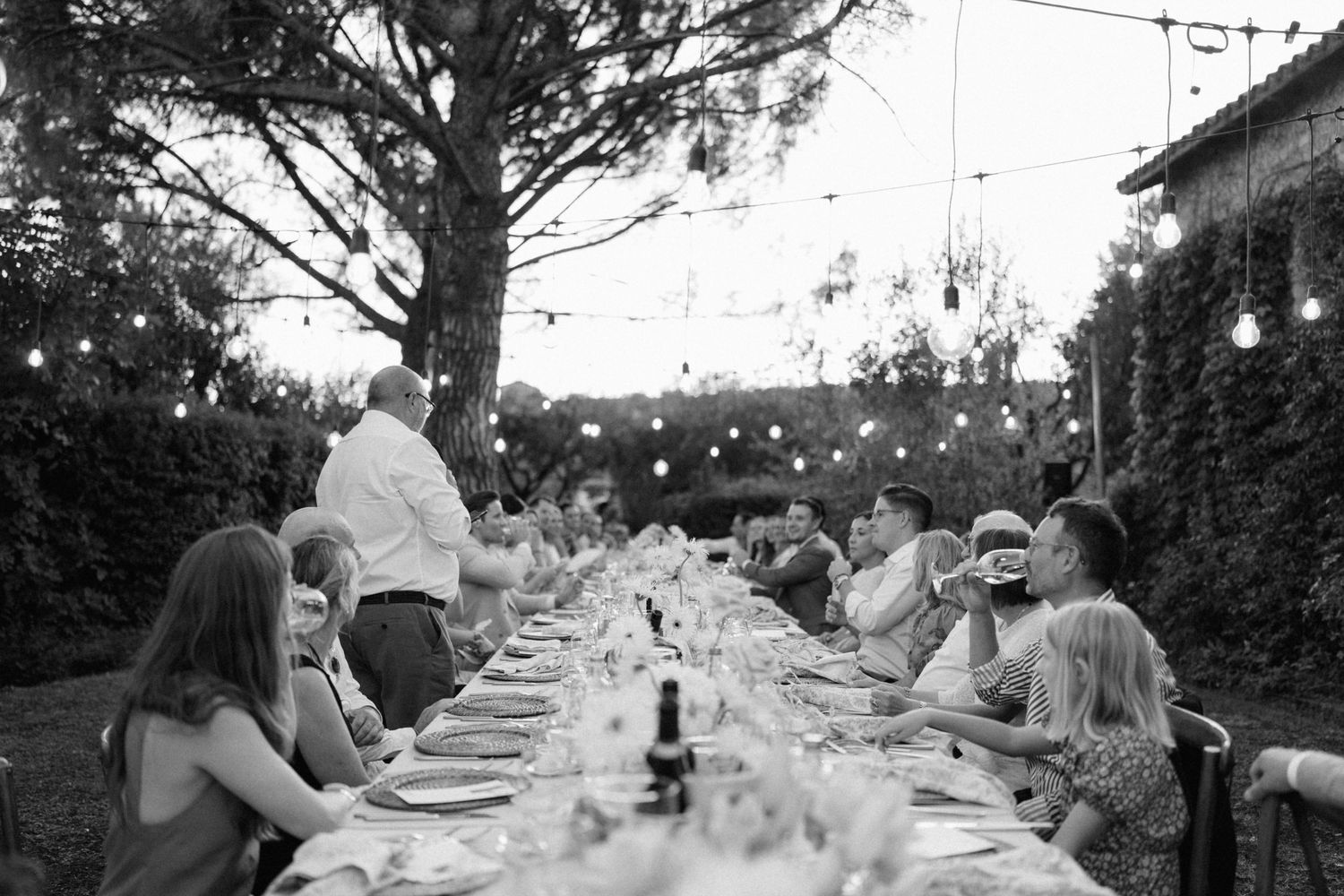 Black and white outdoor dinner party with guests seated at a long table under string lights surrounded by trees.