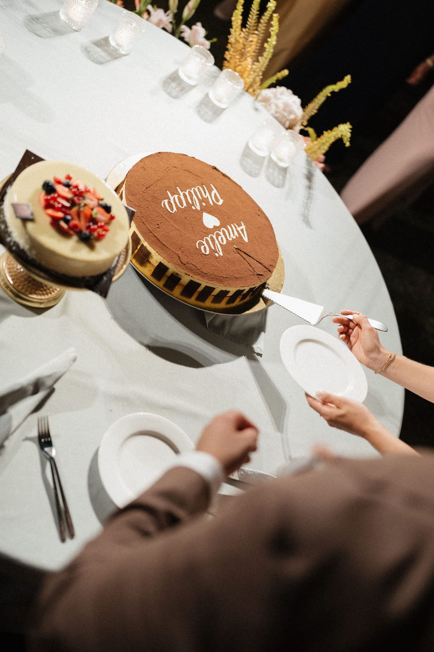 Wedding cake with names Amelie and Philippe on chocolate frosting, being cut at elegant reception table.