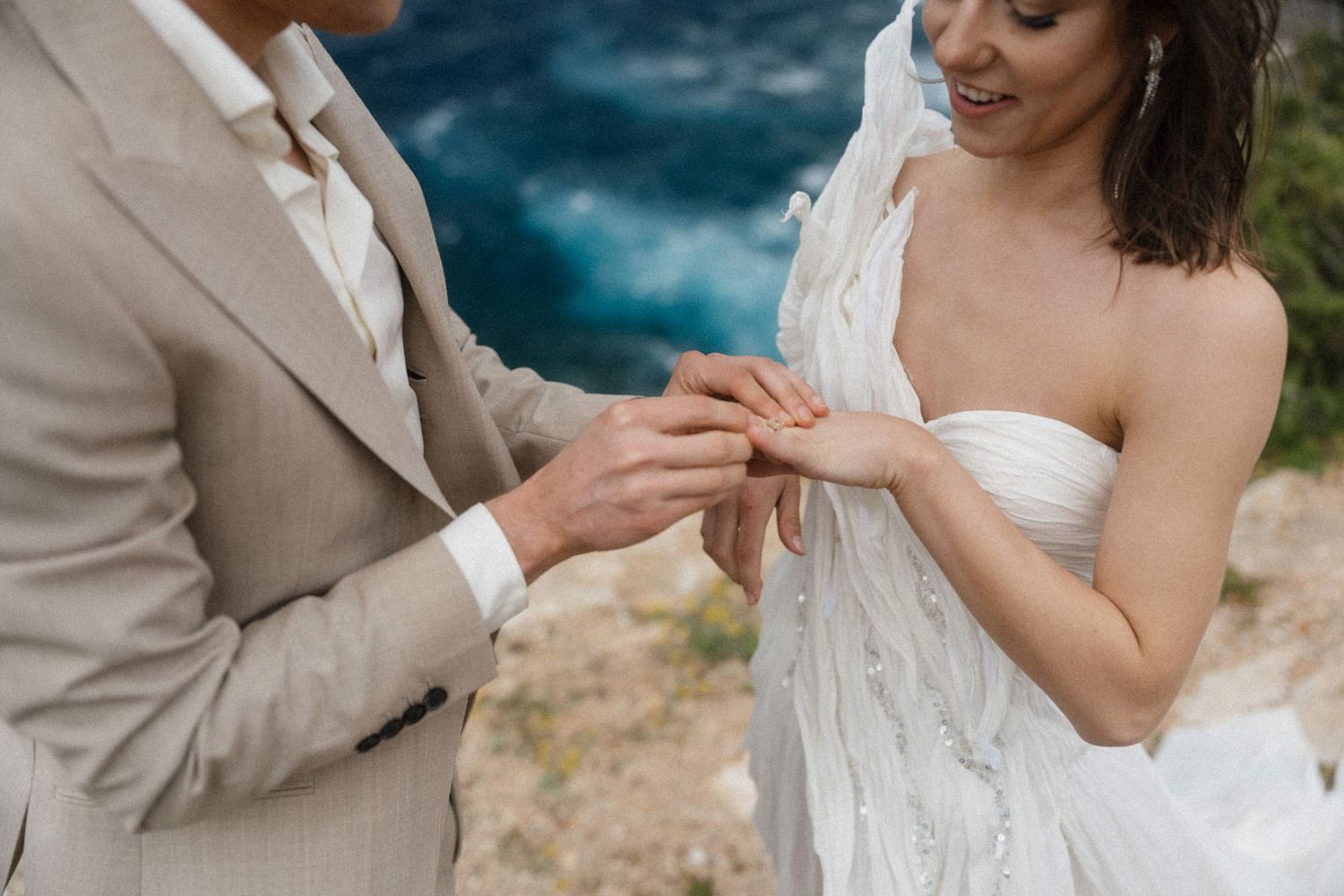 Groom placing a wedding ring on bride's finger during outdoor ceremony with turquoise water backdrop.