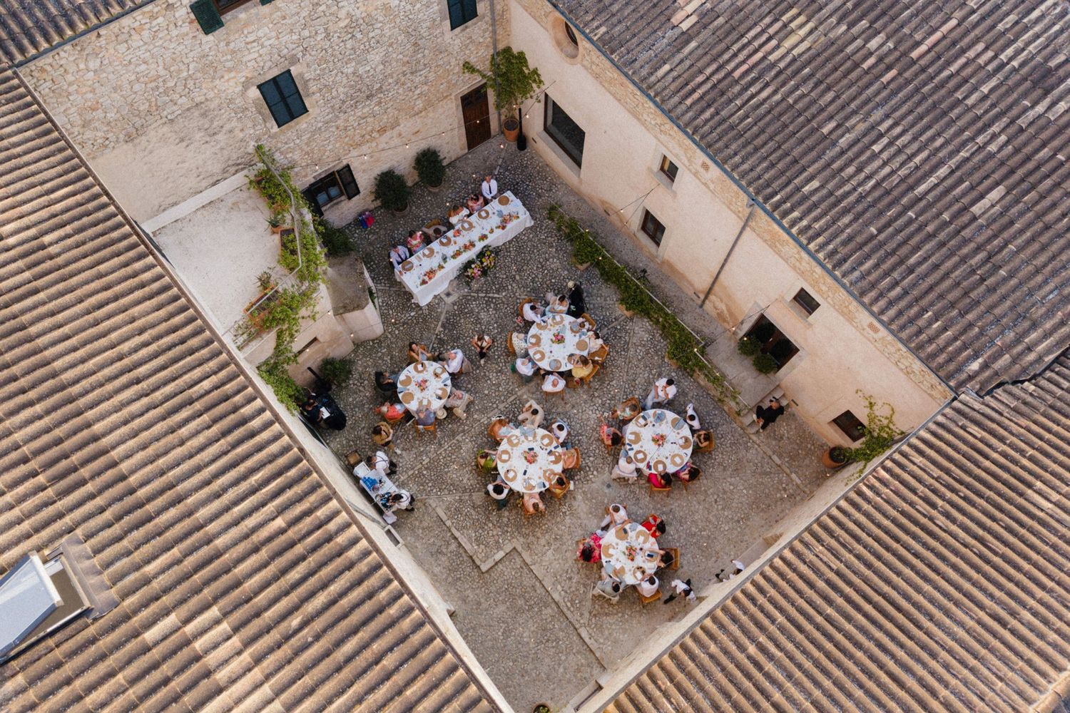 Aerial view of an elegant outdoor courtyard dining event with round tables set for guests in a rustic stone building.