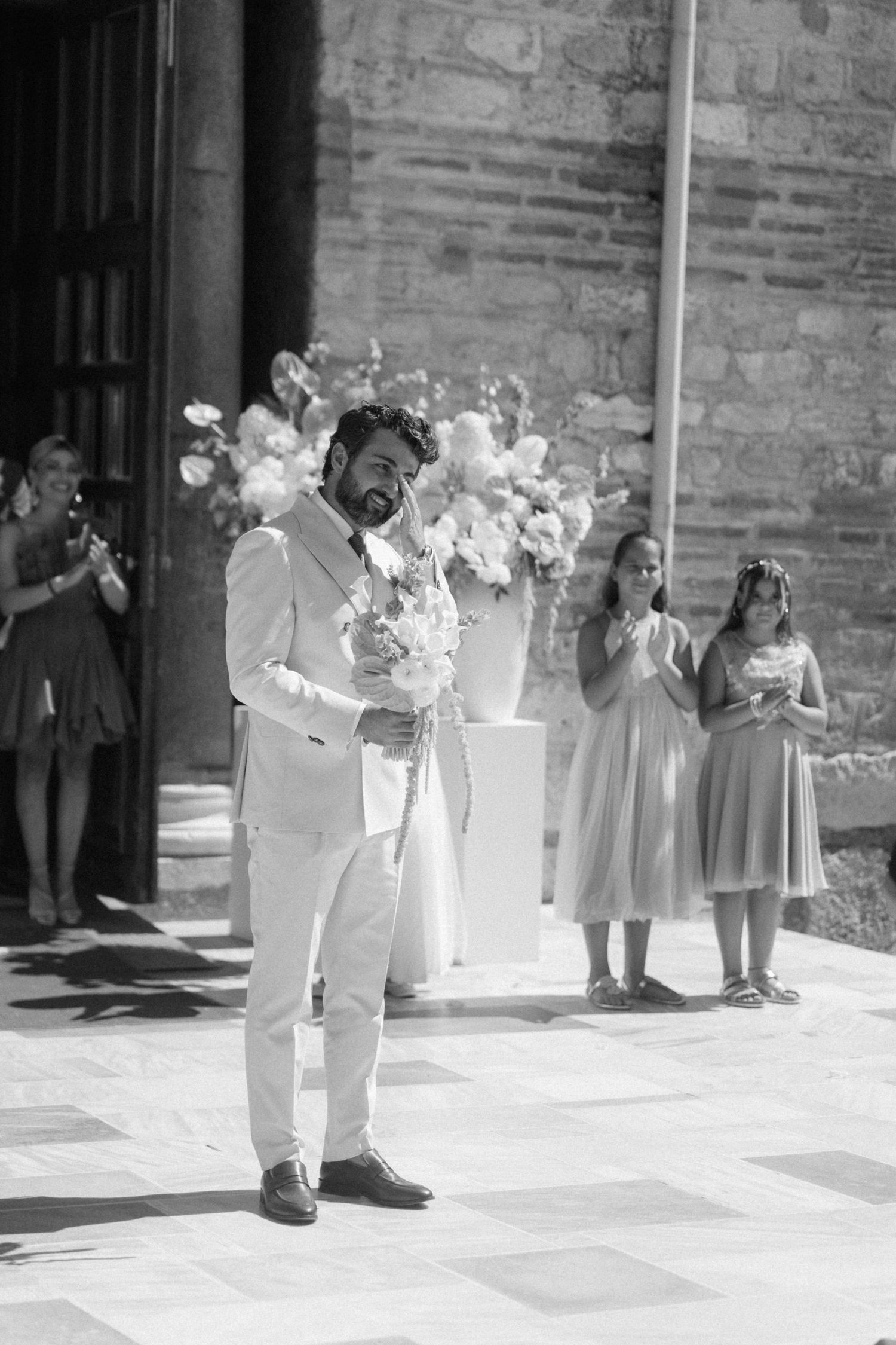 Black and white photo of bearded groom in white suit holding flowers outside decorated church entrance.