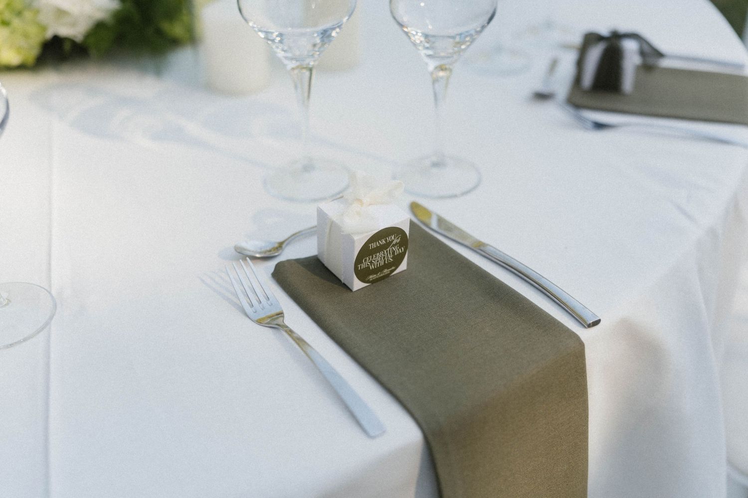 Elegant wedding table setting with white linen, gray napkin, silverware, crystal glasses, and a small favor box.