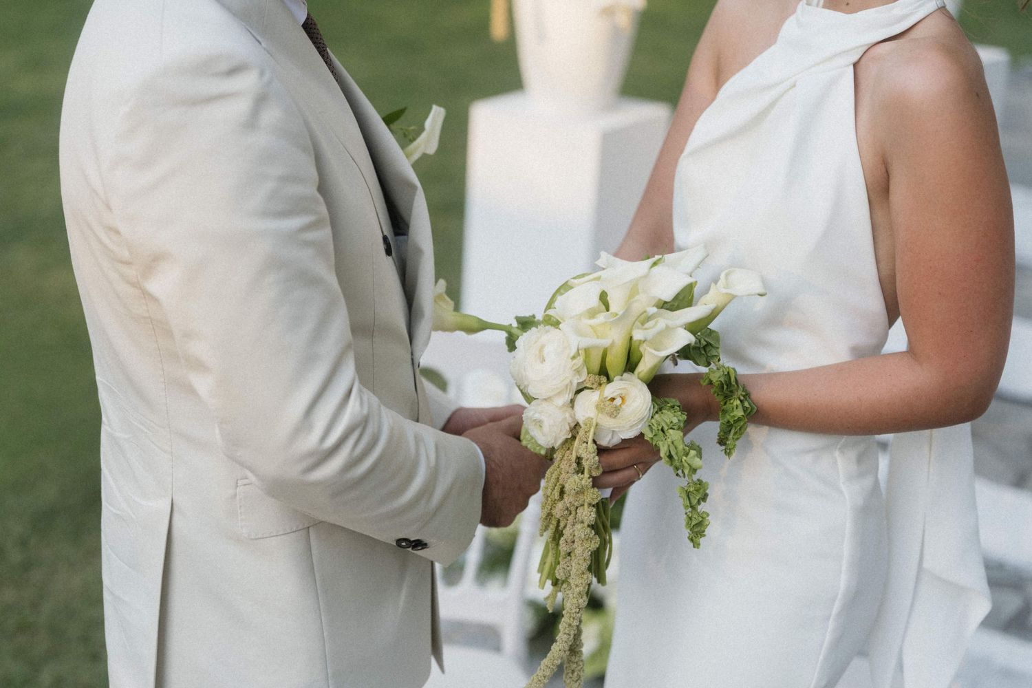 Bride and groom in white wedding attire holding hands, bride carrying elegant white floral bouquet outdoors.