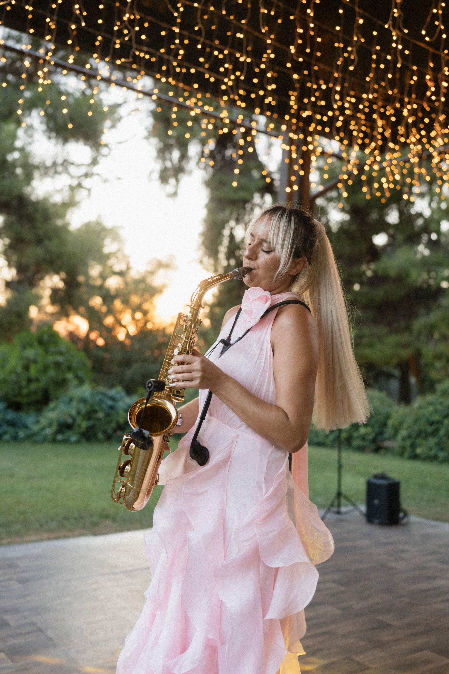 Blonde woman in pink dress playing saxophone outdoors under string lights at sunset garden event.