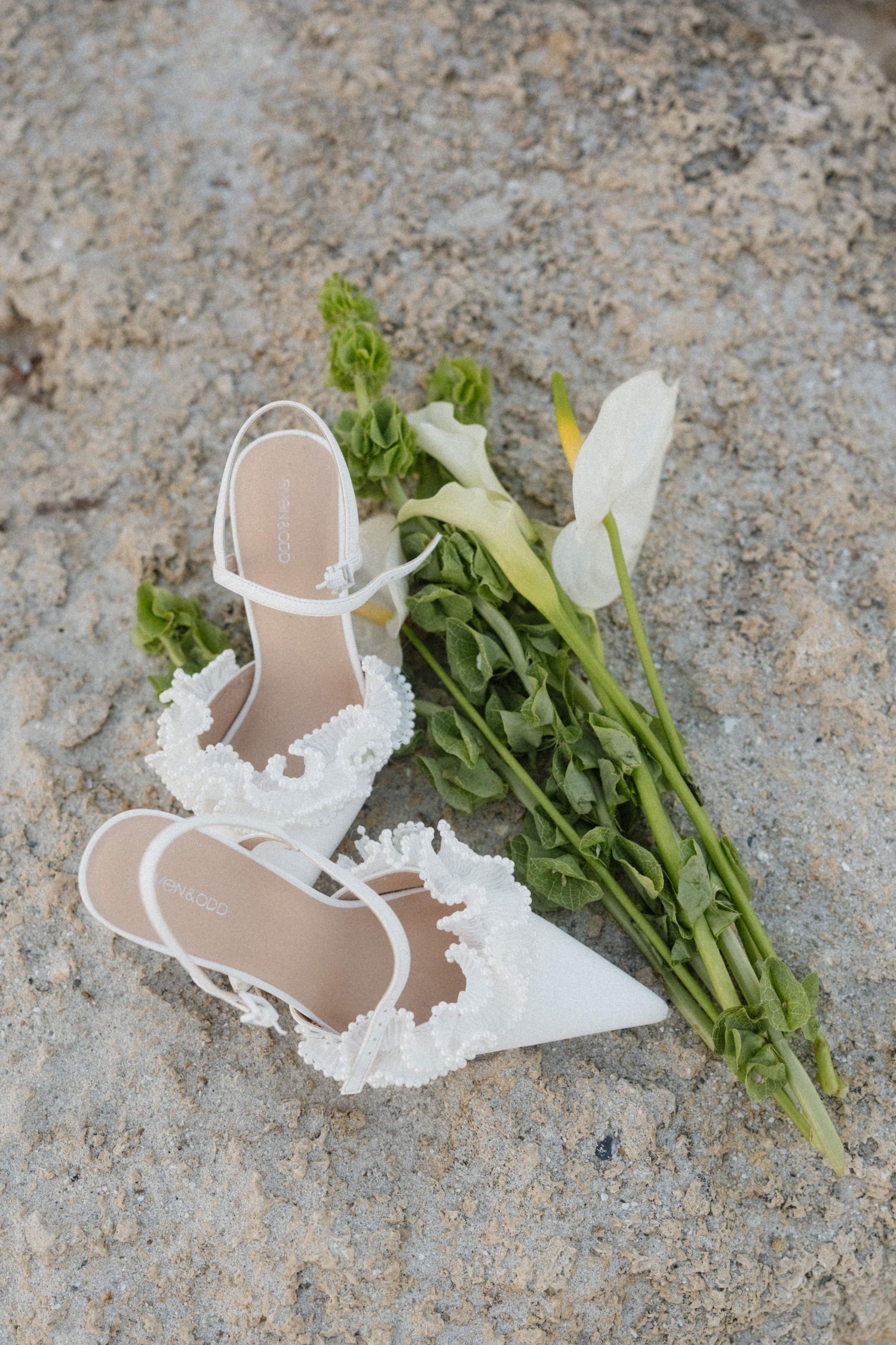 White lace bridal heels and green calla lily bouquet resting on sandy rock surface.