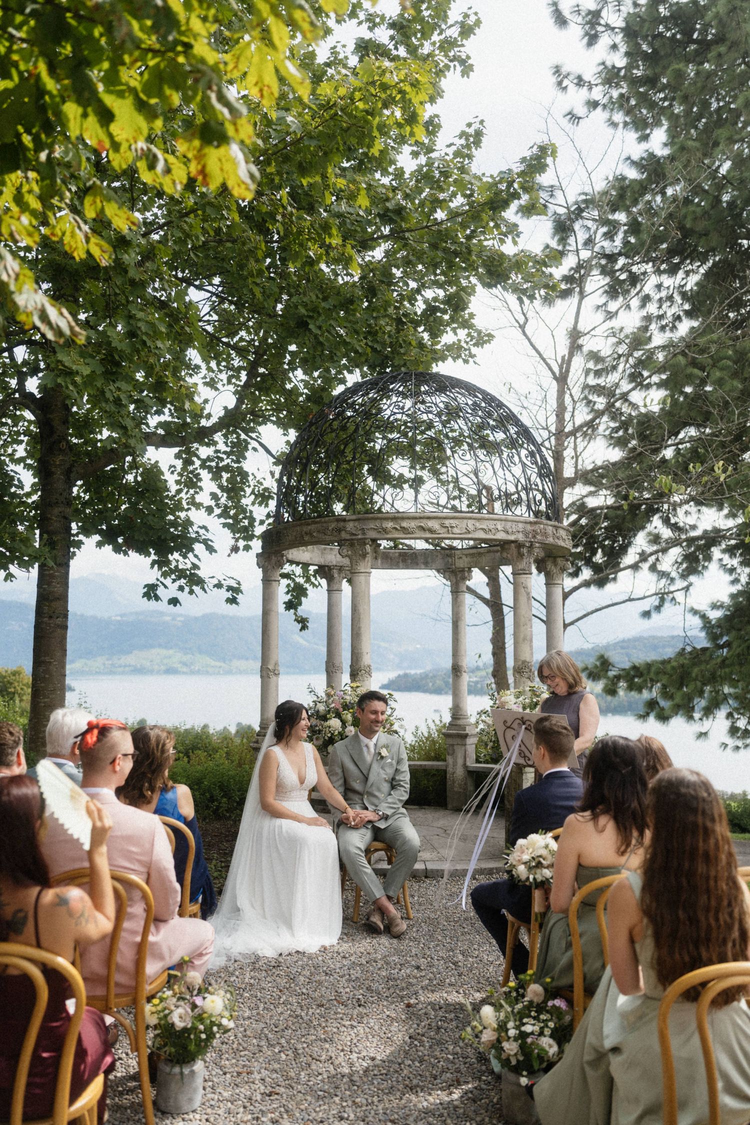 Bride and groom at outdoor wedding ceremony under stone gazebo with lake backdrop.